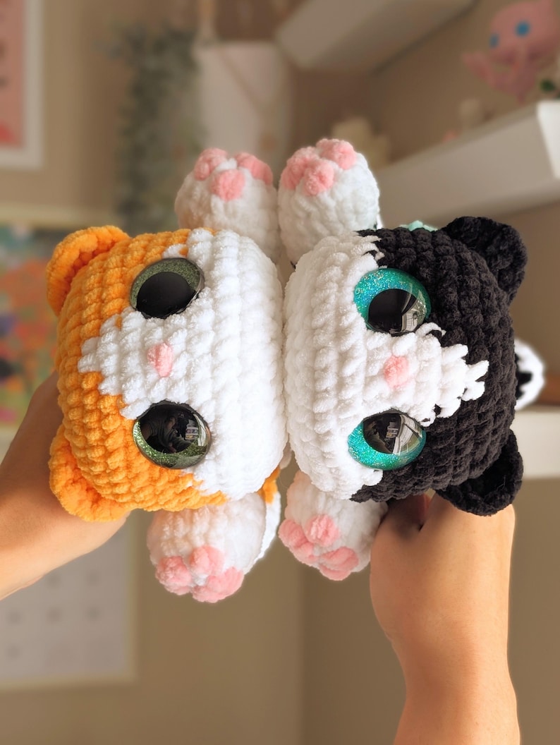 May include: Two crocheted cat plushies, one orange and white, the other black and white. Both have large, glittery eyes and pink paw pads. The plushies are handmade and have a soft, textured appearance.