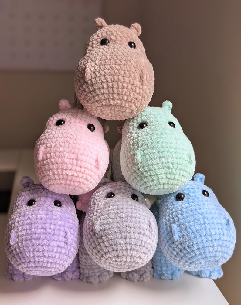 May include: A stack of six crocheted hippopotamus plushies in various pastel colors, including pink, light blue, lavender, and green. The hippos are arranged in a pyramid shape, with the largest hippo at the top.