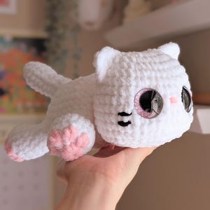 May include: A white crocheted cat plushie with pink accents on the paws and cheeks. The cat has large, glittery eyes and black details for the nose and mouth. The plushie is lying down, with its tail extended.