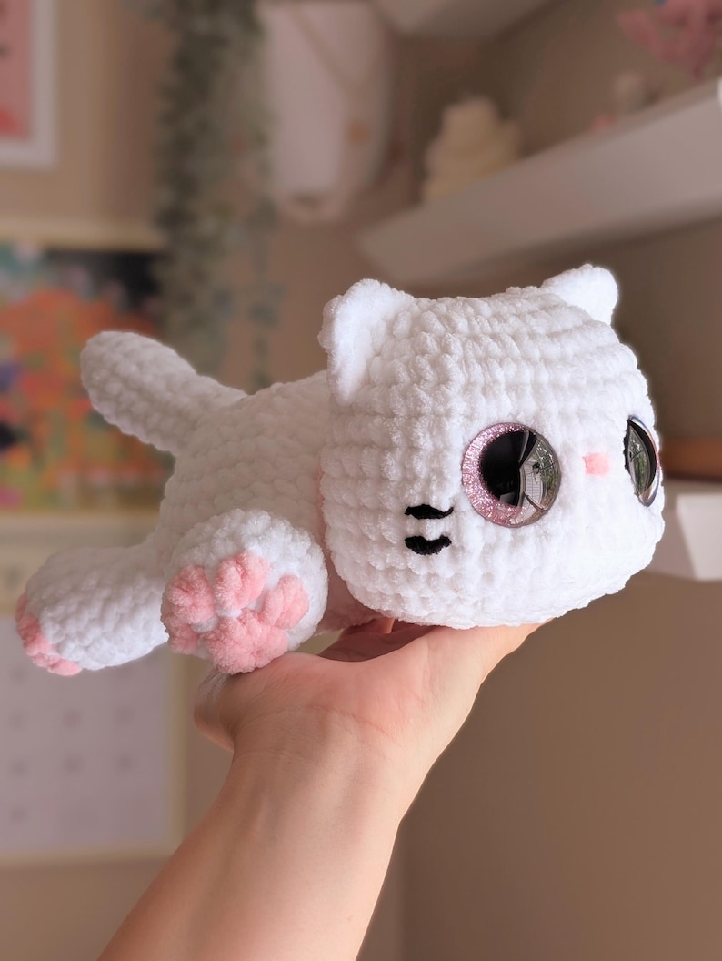 May include: A white crocheted cat plushie with pink accents on the paws and cheeks. The cat has large, glittery eyes and black details for the nose and mouth. The plushie is lying down, with its tail extended.