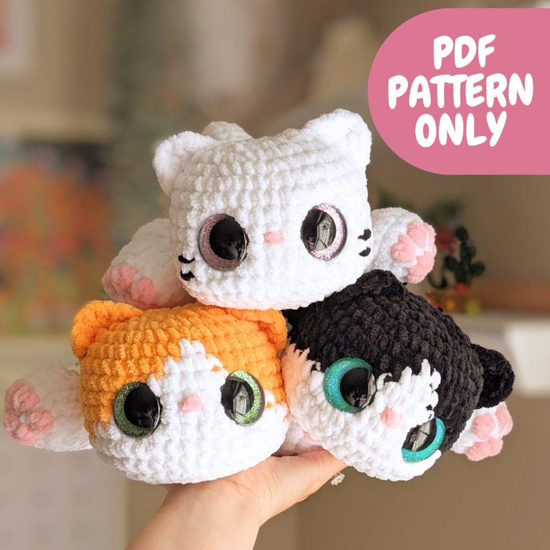 May include: Three handmade crochet cat plushies in white, orange and white, and black and white. Each cat has large, glittery eyes and pink accents. The image includes the text "PDF PATTERN ONLY" in a pink banner.