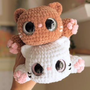 May include: Two handmade crochet cat plushies, one brown and one white, stacked together. The cats have large, reflective eyes, pink paw pads, and small pink accents. The plushies are soft and cuddly.