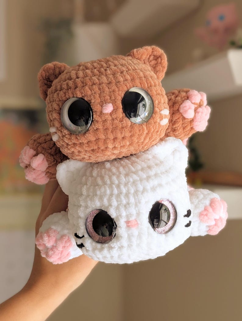 May include: Two handmade crochet cat plushies, one brown and one white, stacked together. The cats have large, reflective eyes, pink paw pads, and small pink accents. The plushies are soft and cuddly.