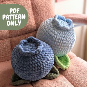 Puede incluir: Dos almohadas de arándano de crochet en tonos azules, con detalles de hojas verdes. Las almohadas están sobre una superficie texturizada rosa. Patrón PDF solamente.