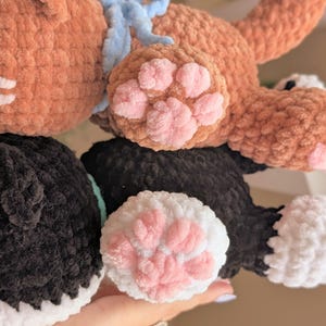 May include: Close-up of handmade crochet cat toys in brown and black. The brown cat has a blue bow and pink paw pads. The black and white cat has pink paw pads. These soft toys are perfect for children.