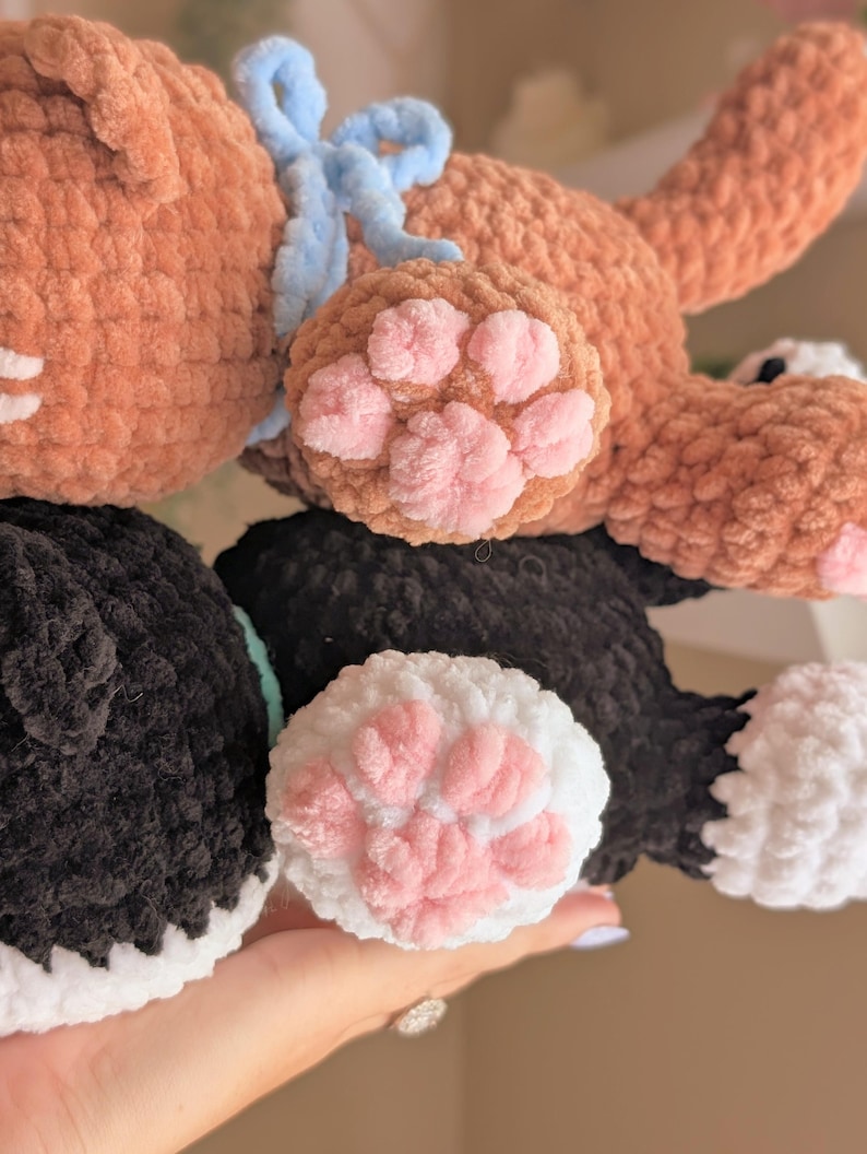 May include: Close-up of handmade crochet cat toys in brown and black. The brown cat has a blue bow and pink paw pads. The black and white cat has pink paw pads. These soft toys are perfect for children.