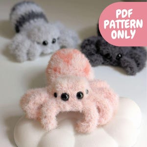 May include: Three fuzzy, crocheted spider figures in light pink, grey, and dark charcoal. Each spider has multiple black bead eyes and soft, textured yarn bodies. A pink graphic overlay in the top right corner contains the text PDF PATTERN ONLY.