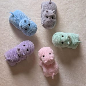 May include: Five crocheted hippopotamus plushies in different colors: light blue, gray, light green, purple, and pink. The plushies are all facing the camera and have black eyes.