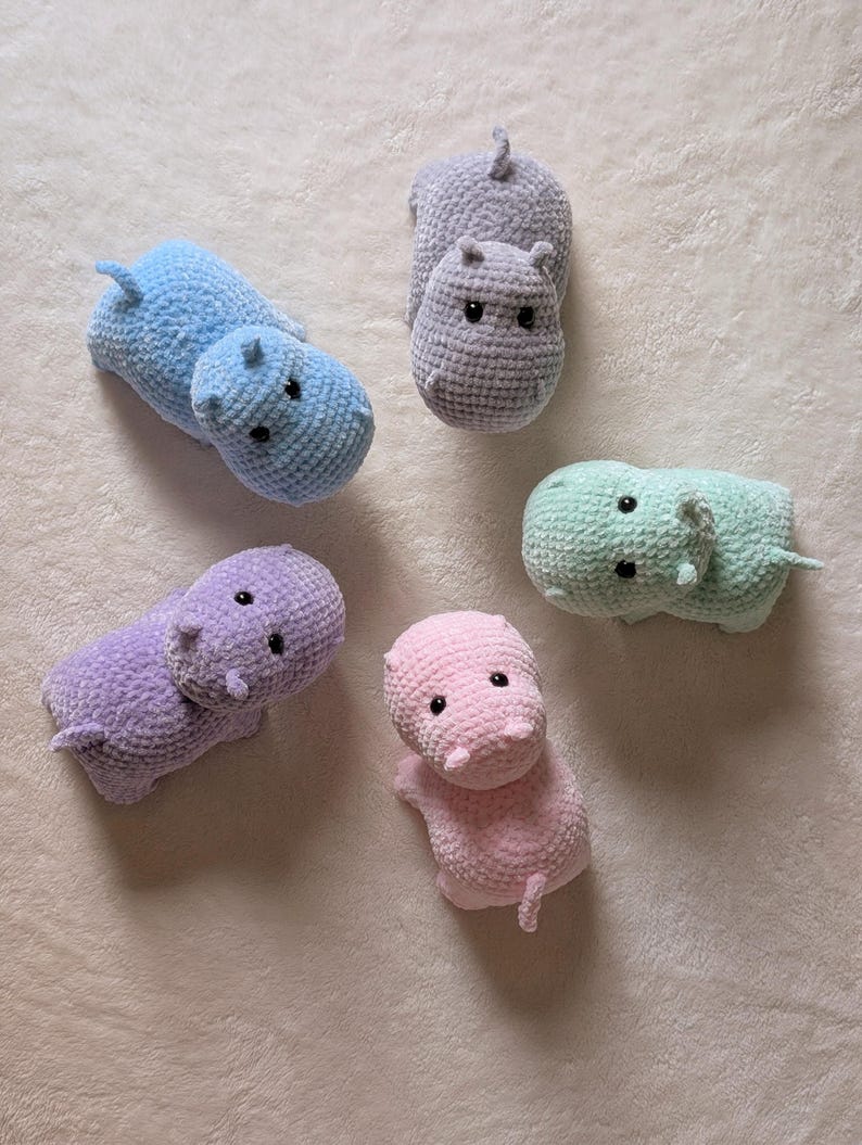 May include: Five crocheted hippopotamus plushies in different colors: light blue, gray, light green, purple, and pink. The plushies are all facing the camera and have black eyes.