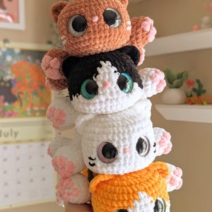 May include: Four handmade crochet cat plushies stacked together. The cats are in shades of orange, black and white, white, and orange and white. Each cat has large, glittery eyes and pink paw pads. The plushies are soft and cuddly.
