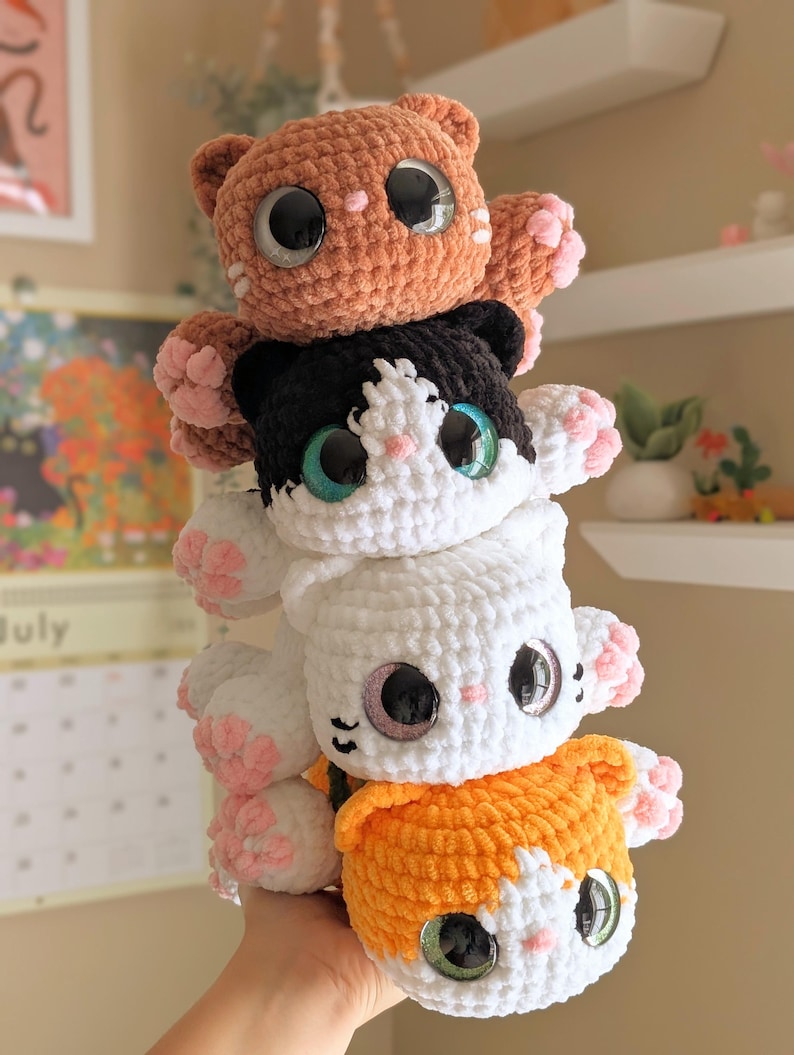 May include: Four handmade crochet cat plushies stacked together. The cats are in shades of orange, black and white, white, and orange and white. Each cat has large, glittery eyes and pink paw pads. The plushies are soft and cuddly.
