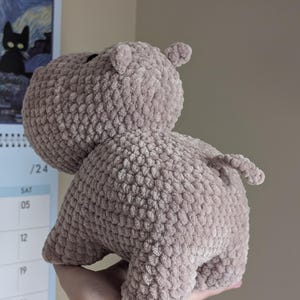 May include: A crocheted plush toy hippopotamus in a light brown color. The hippo is standing on all fours with its tail curled up.