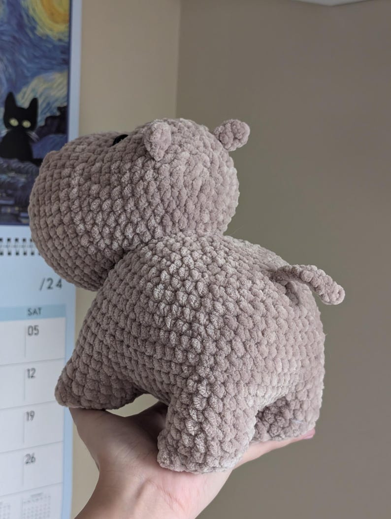 May include: A crocheted plush toy hippopotamus in a light brown color. The hippo is standing on all fours with its tail curled up.