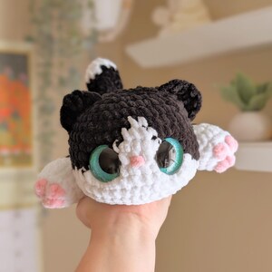 May include: A handmade crochet cat plushie with black and white fur, large teal eyes, and pink paw pads. The cat has a cute, friendly expression and is made with soft yarn. A charming and cuddly stuffed animal.
