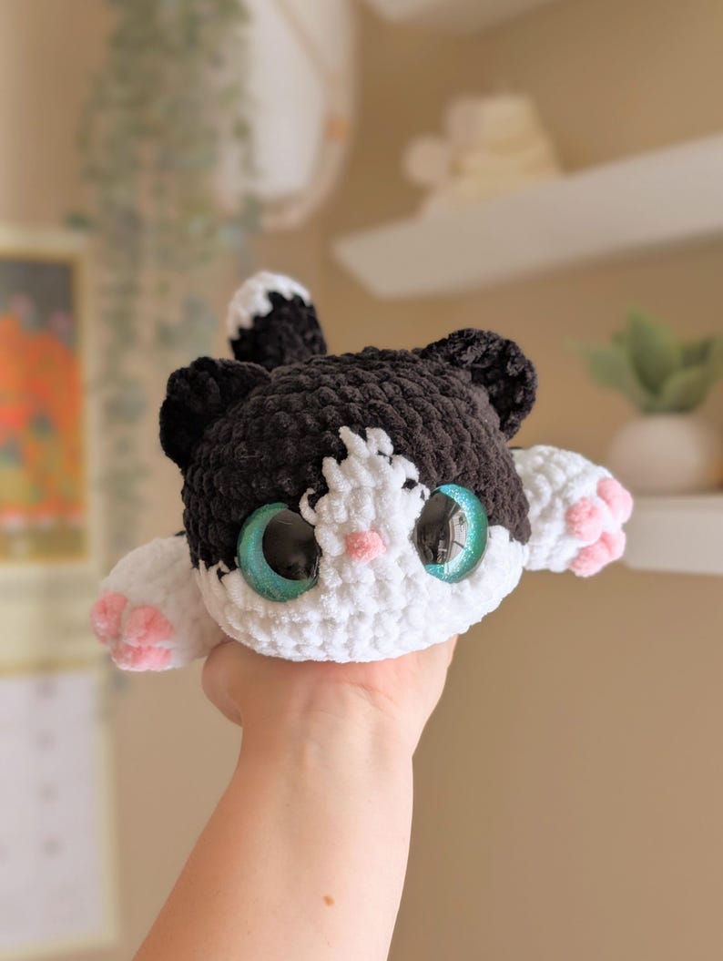 May include: A handmade crochet cat plushie with black and white fur, large teal eyes, and pink paw pads. The cat has a cute, friendly expression and is made with soft yarn. A charming and cuddly stuffed animal.