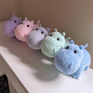 May include: Five crocheted hippopotamus plushies in a row on a white surface. The hippos are in shades of purple, pink, light blue, green, and blue.