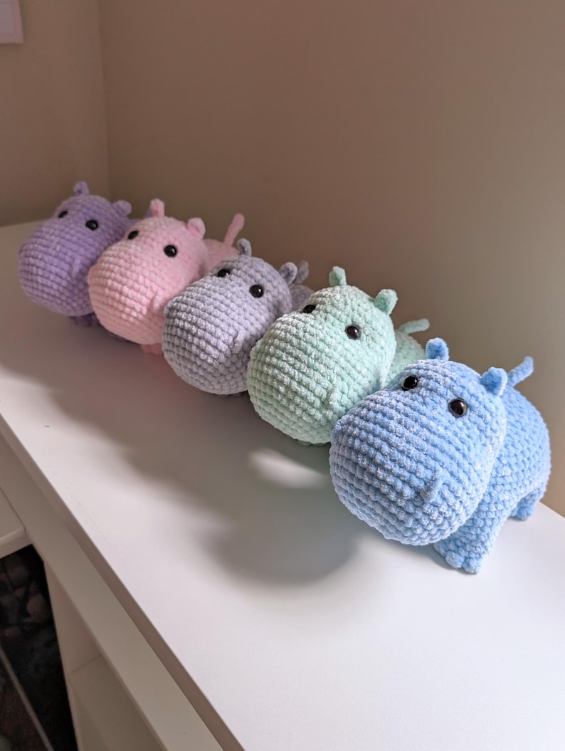 May include: Five crocheted hippopotamus plushies in a row on a white surface. The hippos are in shades of purple, pink, light blue, green, and blue.