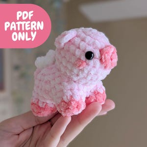 May include: A pink crocheted pig plush toy with black eyes. The toy is being held in a hand. The text "PDF PATTERN ONLY" is in a pink circle in the background.