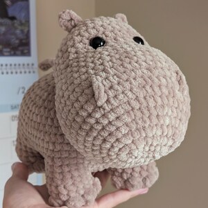 May include: A crocheted plush hippopotamus toy in a light brown color with black eyes. The hippopotamus is standing on its four legs and is being held in a person's hand.