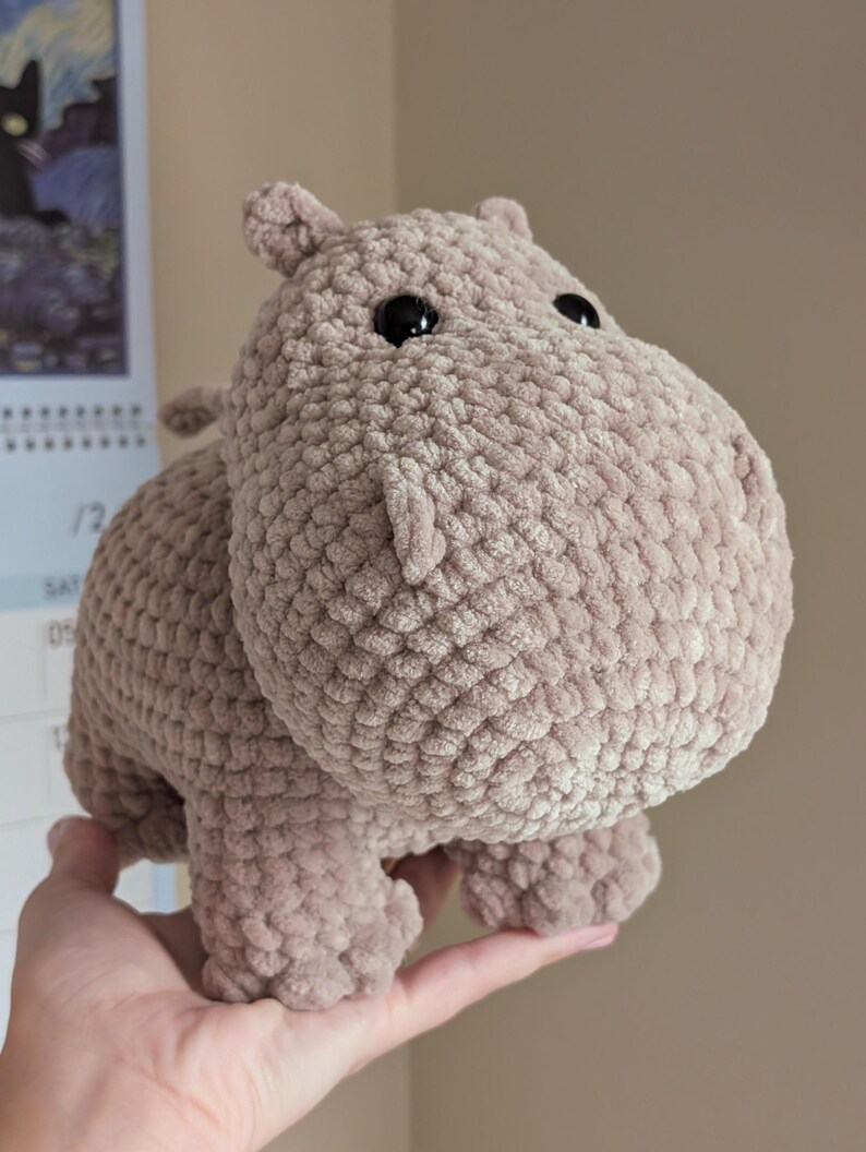 May include: A crocheted plush hippopotamus toy in a light brown color with black eyes. The hippopotamus is standing on its four legs and is being held in a person's hand.