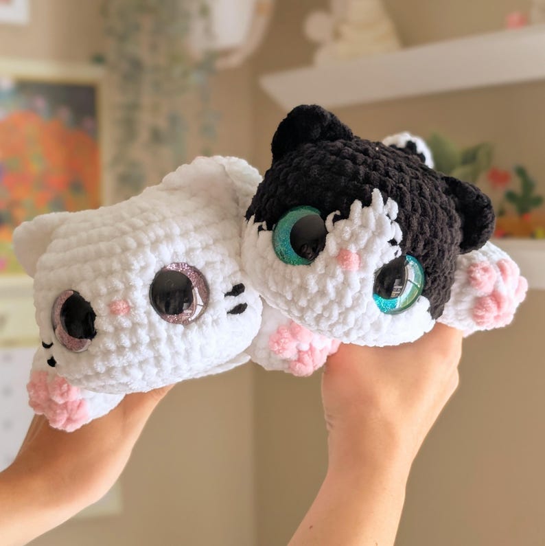 May include: Two crocheted cat plushies. One is white, the other is black and white. Both have large, glittery eyes and pink accents on their cheeks and paws. The plushies are soft and cuddly, ideal as a child's toy or a decorative item.
