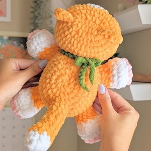 May include: A handmade orange crochet fox plush toy with white paws and a white patch on its head. The fox has a green bow tie and pink accents on its paws. The plush toy is soft and fluffy.