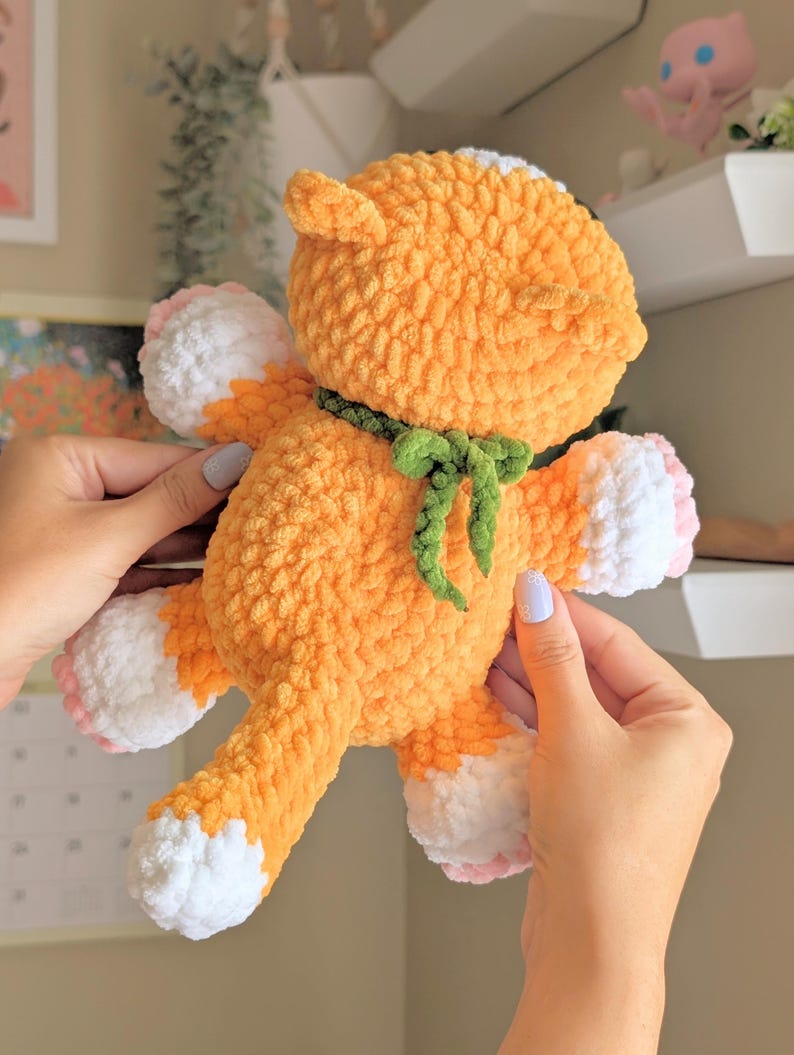 May include: A handmade orange crochet fox plush toy with white paws and a white patch on its head. The fox has a green bow tie and pink accents on its paws. The plush toy is soft and fluffy.