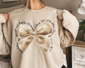 May include: A beige sweatshirt with a brown watercolour bow and the text "She is clothed with strength and dignity. She laughs without fear of the future. Proverbs 31:25" printed on it.
