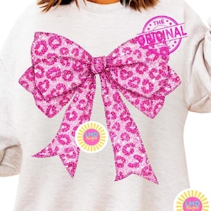 May include: A light gray sweatshirt featuring a large, pink leopard print bow design. The bow has a glittery texture and is the focal point of the shirt. The text "THE ORIGINAL" is stamped on the top right of the bow.