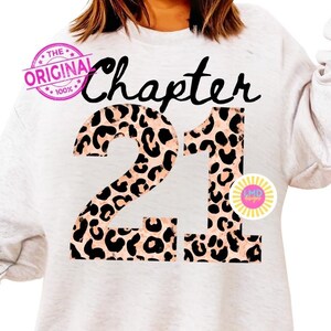 May include: A light gray sweatshirt with the words "Chapter 21" in black script and leopard print. The words "The Original 100%" are printed in a pink circle on the upper left. A yellow and pink sticker is on the right.