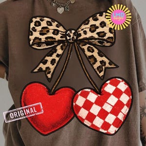 May include: Brown t-shirt featuring a leopard print bow above two heart-shaped cherries. One heart is solid red, and the other has a red and white checkered pattern. The word "ORIGINAL" is printed on a small rectangle.