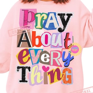 May include: A light pink sweatshirt with the words "Pray About Every Thing" in a colorful, collage-style design. The text is in various fonts and colors, including pink, black, yellow, and blue. The sweatshirt has long sleeves.