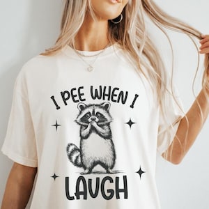 May include: Ivory-colored t-shirt with the text "I Pee When I Laugh" and a graphic of a raccoon. The raccoon is in black and white, with its paws clasped together. Four black star shapes surround the raccoon and text.
