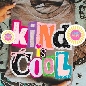 May include: A tan crew neck sweatshirt with the words "Kind is Cool" in a colorful, collage-style design. The text features various fonts and colors, including pink, orange, and blue. The sweatshirt is held by hands.