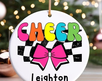 May include: A white ceramic Christmas ornament with a black and white checkered pattern, a pink bow, and the word "CHEER" in rainbow colors. The name "Leighton" is written in black below the bow.
