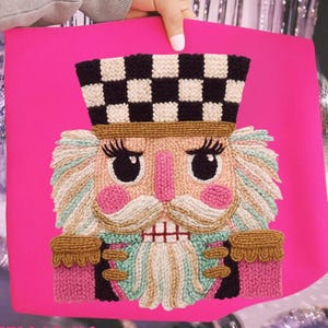 May include: A vibrant pink tote bag with an embroidered nutcracker design. The nutcracker has a black and white chequered hat, pink cheeks, and a white moustache and beard. The word "HELICONIA" is printed at the bottom of the bag.