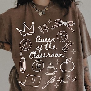 May include: Espresso-colored t-shirt with a white graphic that reads "Queen of the Classroom." The design includes a crown, smiley face, apple, coffee cup, and other school-related icons. The shirt is a casual, comfortable style.