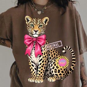 May include: Brown t-shirt featuring a seated leopard illustration with a large pink bow. The leopard has green eyes and a spotted coat. The word "ORIGINAL" is printed on a rectangular label.