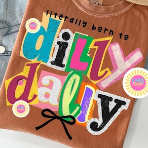 May include: A rust-colored t-shirt with the phrase "literally born to dilly dally" in a colorful, cut-out style. The shirt features a black bow and a round sticker with the text "LMD designs".
