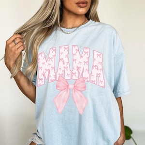 May include: Light blue t-shirt with the word "MAMA" in white letters with a pink bow. The letters are covered in a pattern of small pink bows.