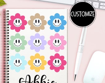 May include: A spiral-bound notebook with a white cover featuring colorful flower designs with smiley faces. The name "Abbie" is written in black script at the bottom. A black circle with the word "CUSTOMIZE" is on the right.