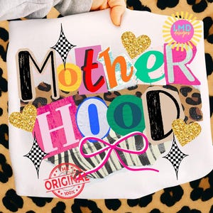 May include: White t-shirt with the words "MOTHERHOOD" in a colorful, collage-style design. The design includes leopard print, zebra stripes, glitter hearts, and a pink bow. The shirt also features a "THE ORIGINAL 100%" stamp.