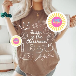 May include: Brown t-shirt with the phrase "Queen of the Classroom" and various school-related doodles in white. The doodles include a crown, apple, laptop, and coffee cup. The shirt is a neutral color.