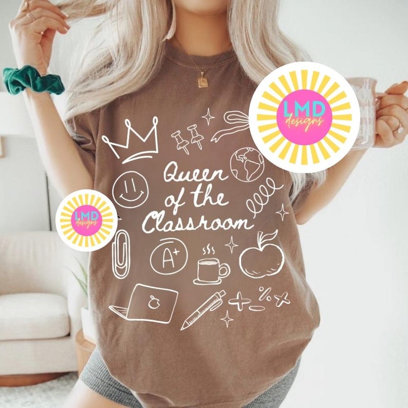 May include: Brown t-shirt with the phrase "Queen of the Classroom" and various school-related doodles in white. The doodles include a crown, apple, laptop, and coffee cup. The shirt is a neutral color.