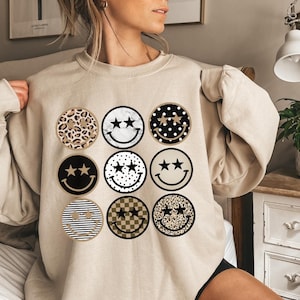 May include: A beige sweatshirt with a graphic print of nine smiley faces. Each smiley face has a different pattern, including leopard print, marble, polka dots, checkered, and stripes. The smiley faces have two gold stars for eyes.
