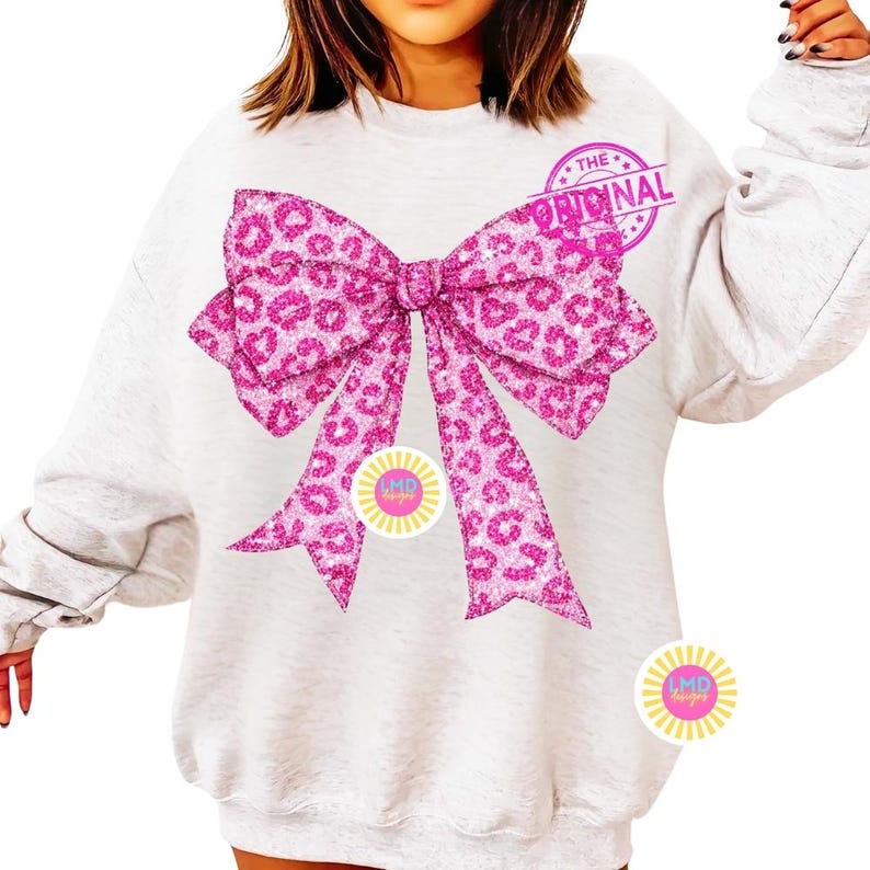 May include: A light gray sweatshirt featuring a large, pink leopard print bow design. The bow has a glittery texture and is the focal point of the shirt. The text "THE ORIGINAL" is stamped on the top right of the bow.