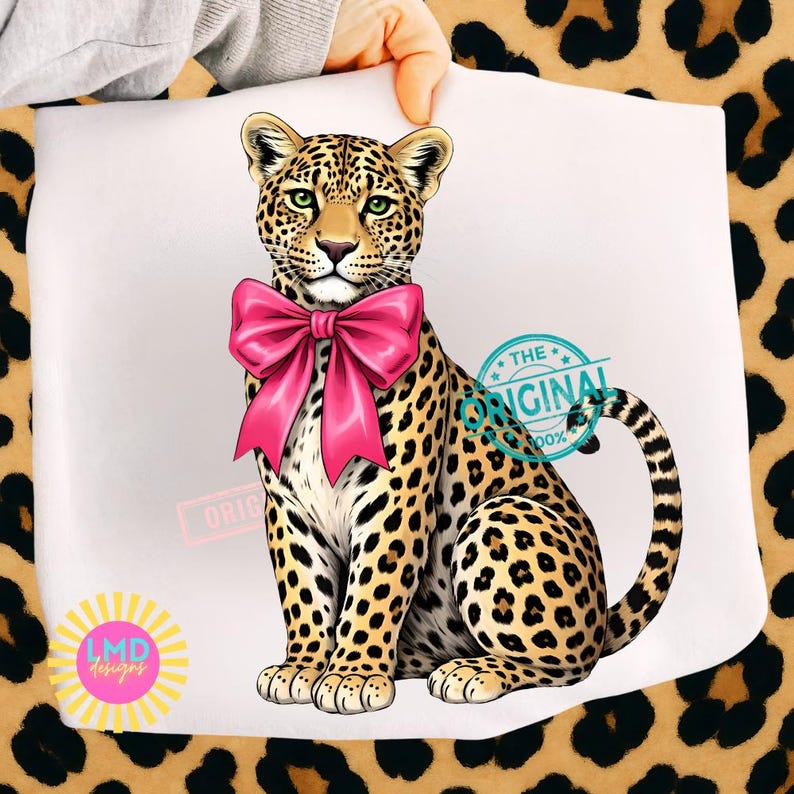 May include: A digitally illustrated leopard with a large pink bow around its neck. The leopard has a spotted coat, green eyes, and is sitting upright. The image includes the text "THE ORIGINAL" and "LMD designs".