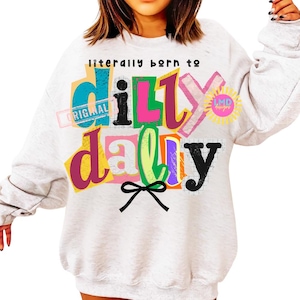 May include: A light gray sweatshirt with the phrase "literally born to dilly dally" in colorful, layered text. The design includes a bow and a sun graphic. The word "original" is also visible.