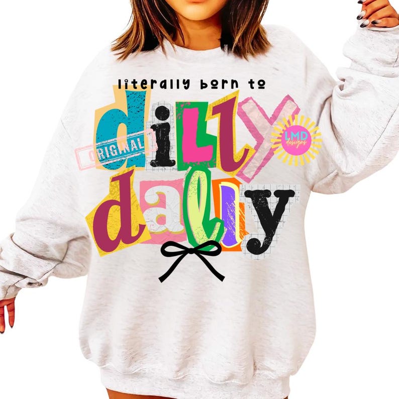 May include: A light gray sweatshirt with the phrase "literally born to dilly dally" in colorful, layered text. The design includes a bow and a sun graphic. The word "original" is also visible.