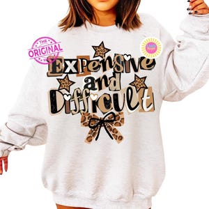 Pu&ograve; includere: Felpa grigio chiaro con la scritta "Expensive and Difficult" in un mix di font e colori. Il design include stelle con stampa leopardata e un fiocco. Il testo &egrave; in nero, marrone e beige.